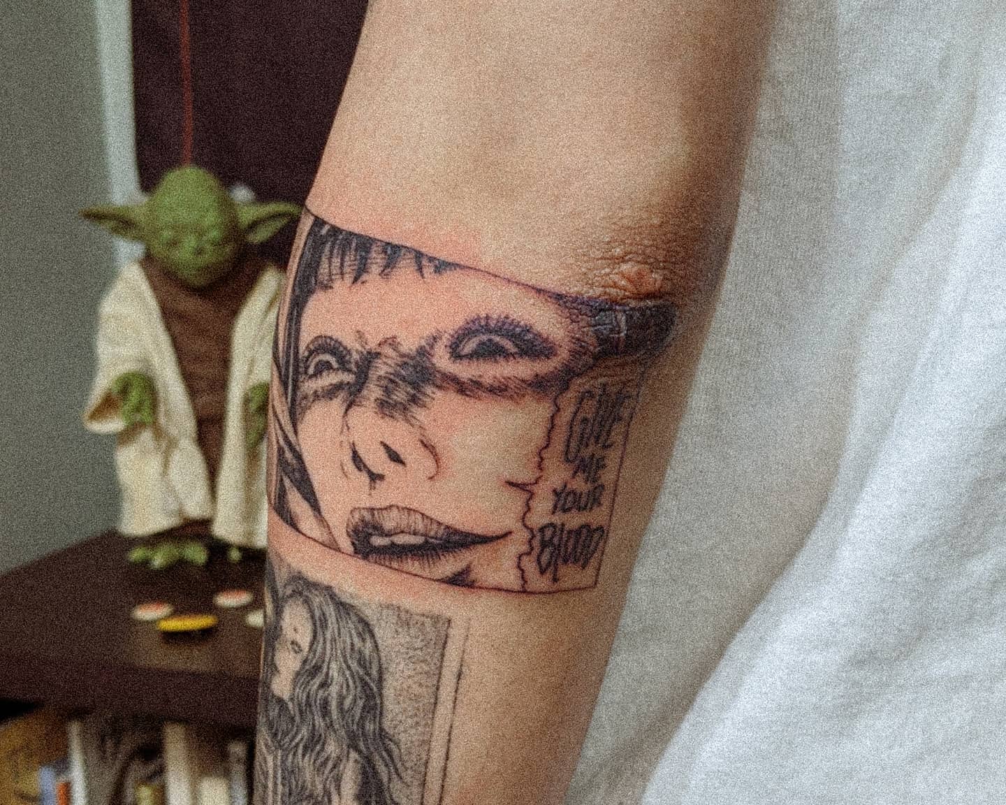 Forearm tattoo of a grinning Junji Ito character with text reading 'Give me your blood' in a creepy panel
