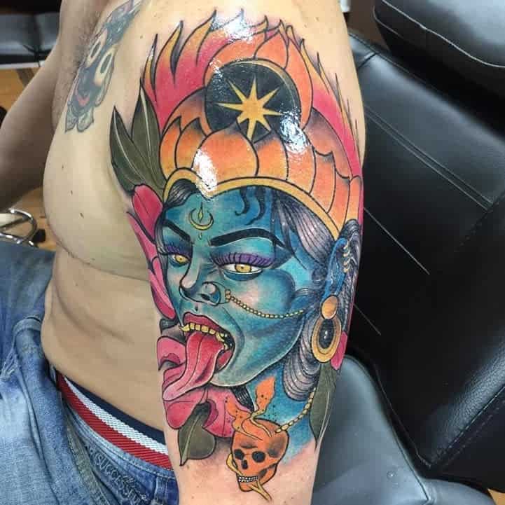 Vibrant Kali tattoo, featuring the blue-skinned goddess with a fiery crown, extended tongue, pink flowers and green leaves and a skull pendant
