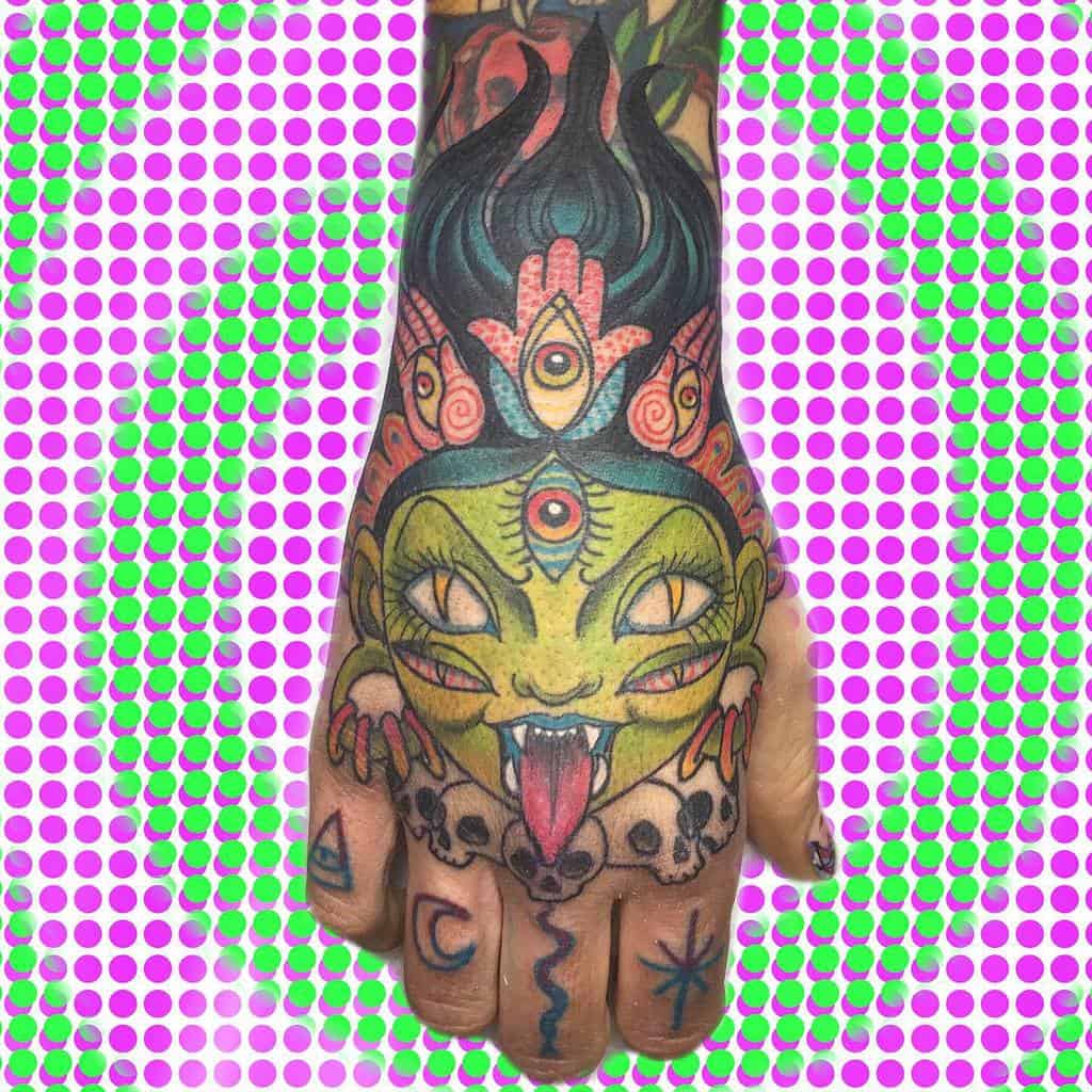 Colorful Kali-inspired hand tattoo featuring a green-faced deity with a third eye, fiery crown, extended tongue, and skull necklace
