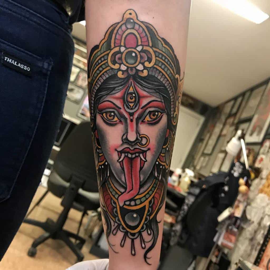 Intricate Kali tattoo on the forearm, featuring the fierce goddess with a third eye, ornate headdress, extended tongue, and bold shading