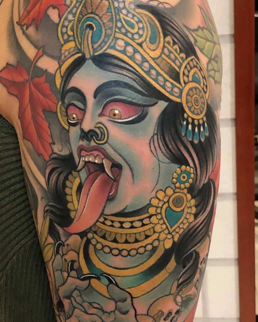 Vibrant Kali tattoo, depicting the fierce blue-skinned goddess with glowing eyes, fangs, extended tongue, ornate jewelry, and skulls