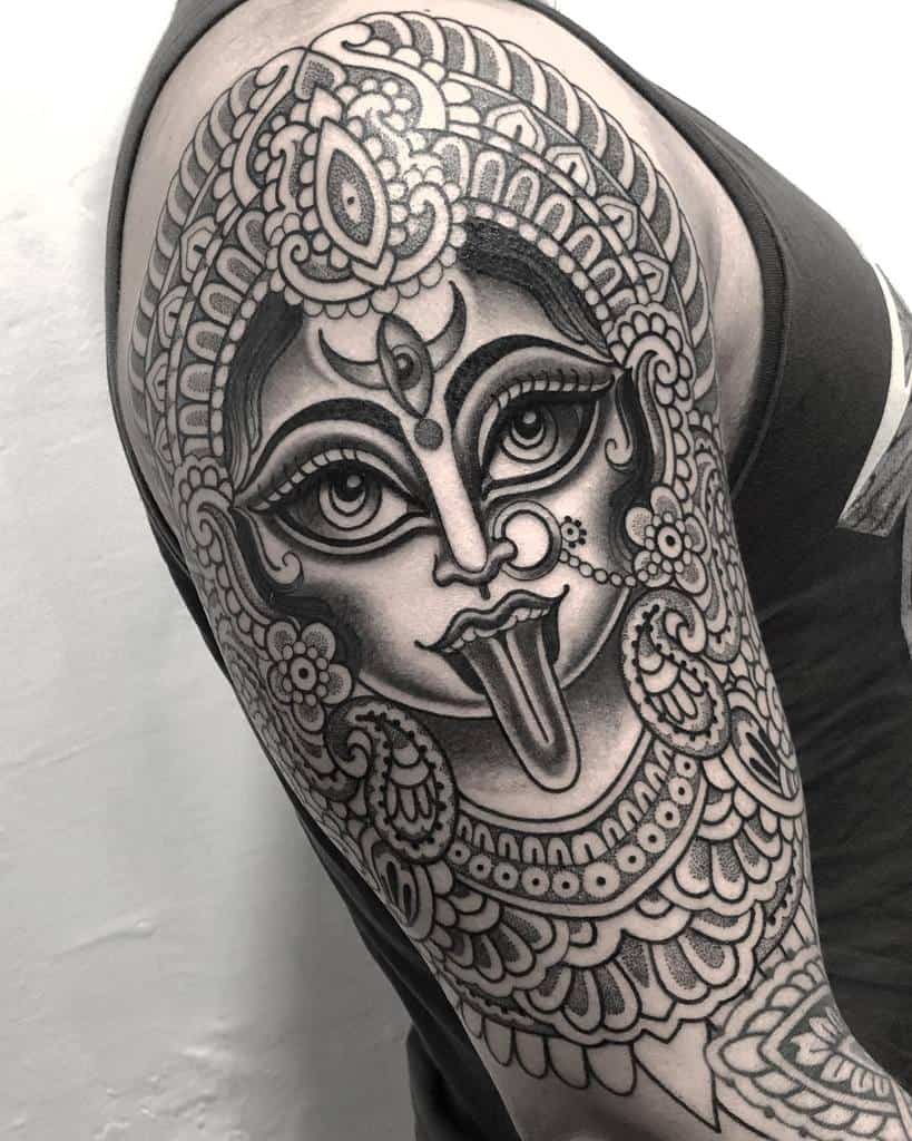 Intricate black and grey Kali tattoo, featuring the goddess with a third eye, ornate headdress, extended tongue, and detailed jewelry