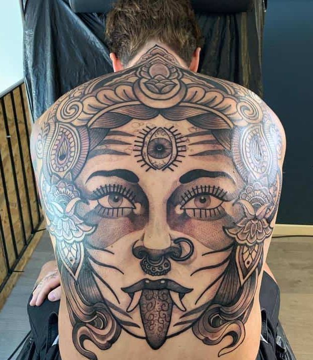 Black and grey tattoo of Kali, featuring an ornate headdress, a third eye, detailed floral patterns, and an extended tongue with sacred script