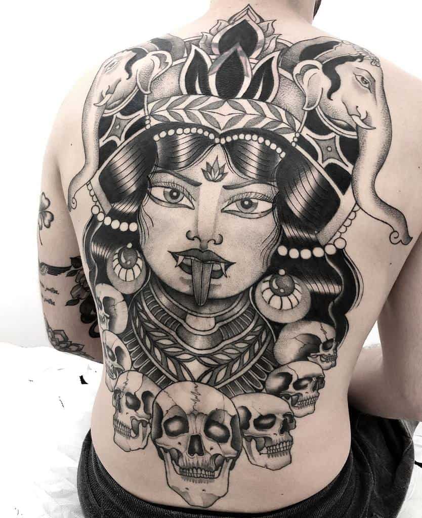 Black and grey tattoo of goddess Kali, featuring an ornate headdress, third eye, extended tongue, skull necklace, and elephant motifs