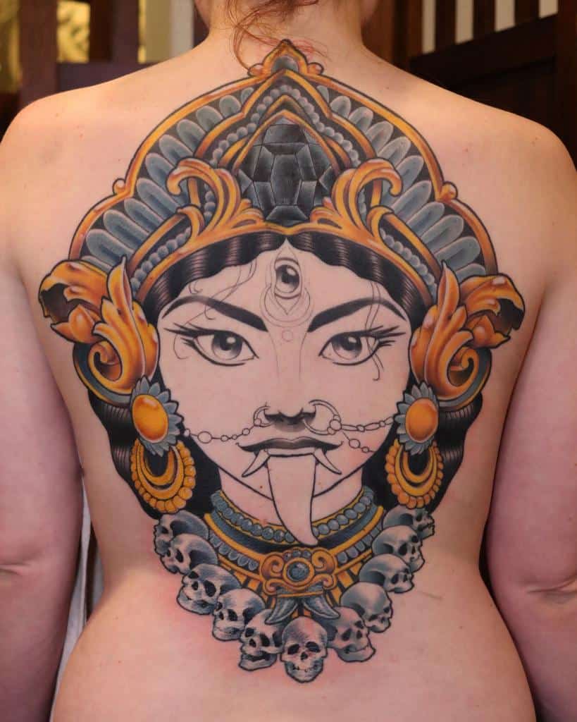 Kali tattoo featuring an elaborate golden and black headdress, a third eye, extended tongue, and a detailed skull necklace