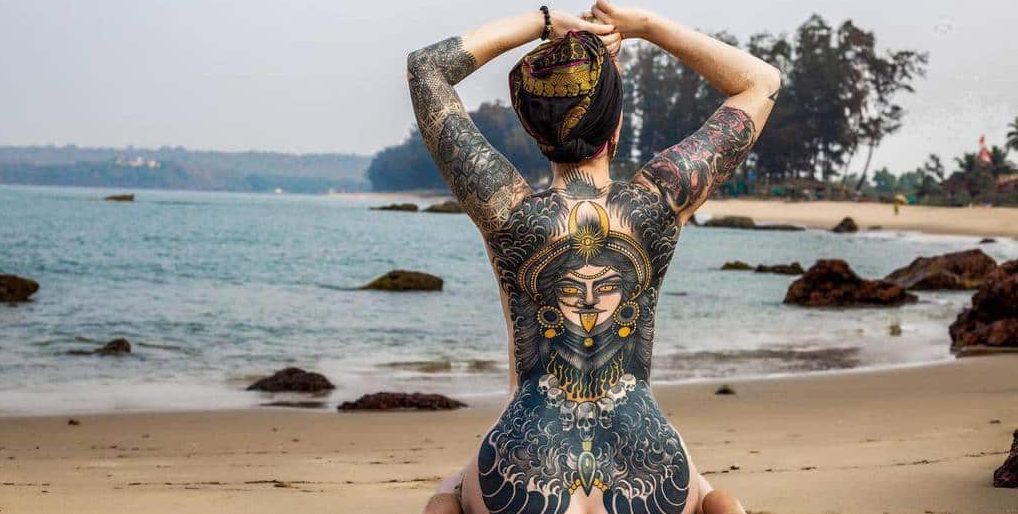 Kali tattoo featuring an ornate golden and black headdress, intense gaze, extended tongue, intricate jewelry, and skulls, blending seamlessly with flowing pattern