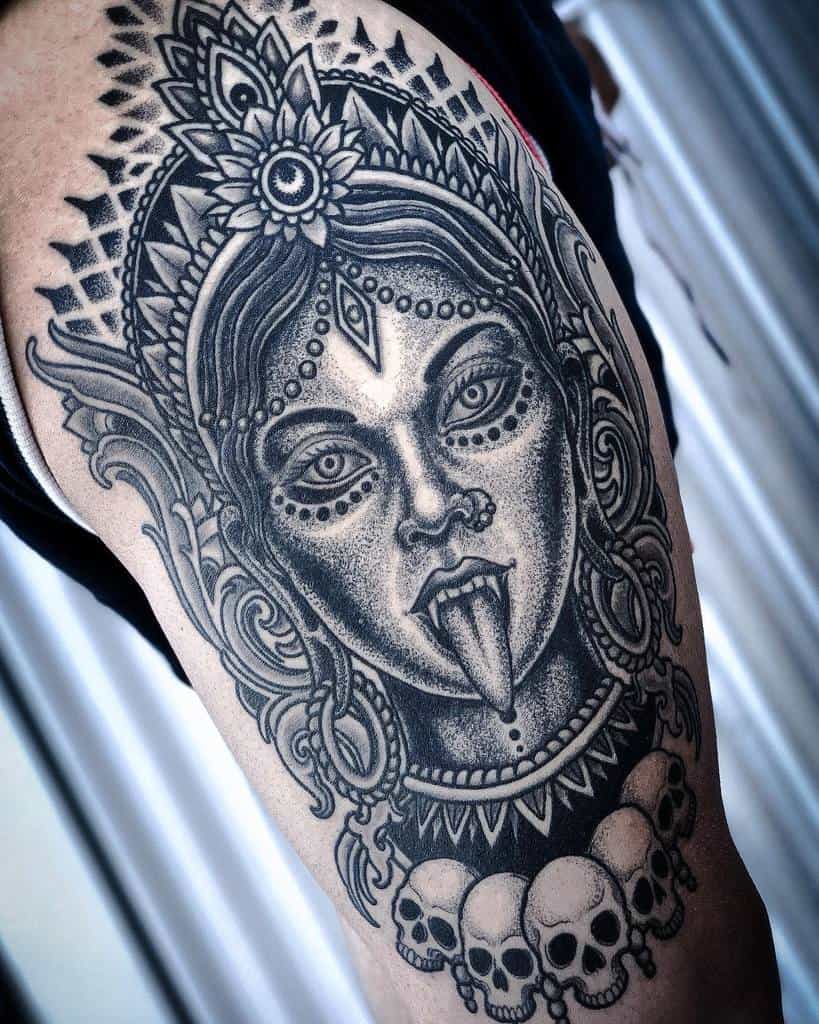 Detailed black and grey Kali tattoo, featuring an ornate headdress, third eye, extended tongue, and a skull necklace with bold dotwork shading