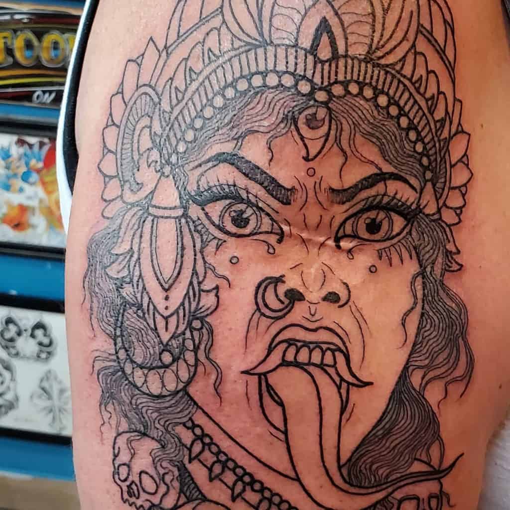 Black ink outline tattoo of goddess Kali, featuring an ornate headdress, fierce expression, extended tongue, and skull-adorned jewelry