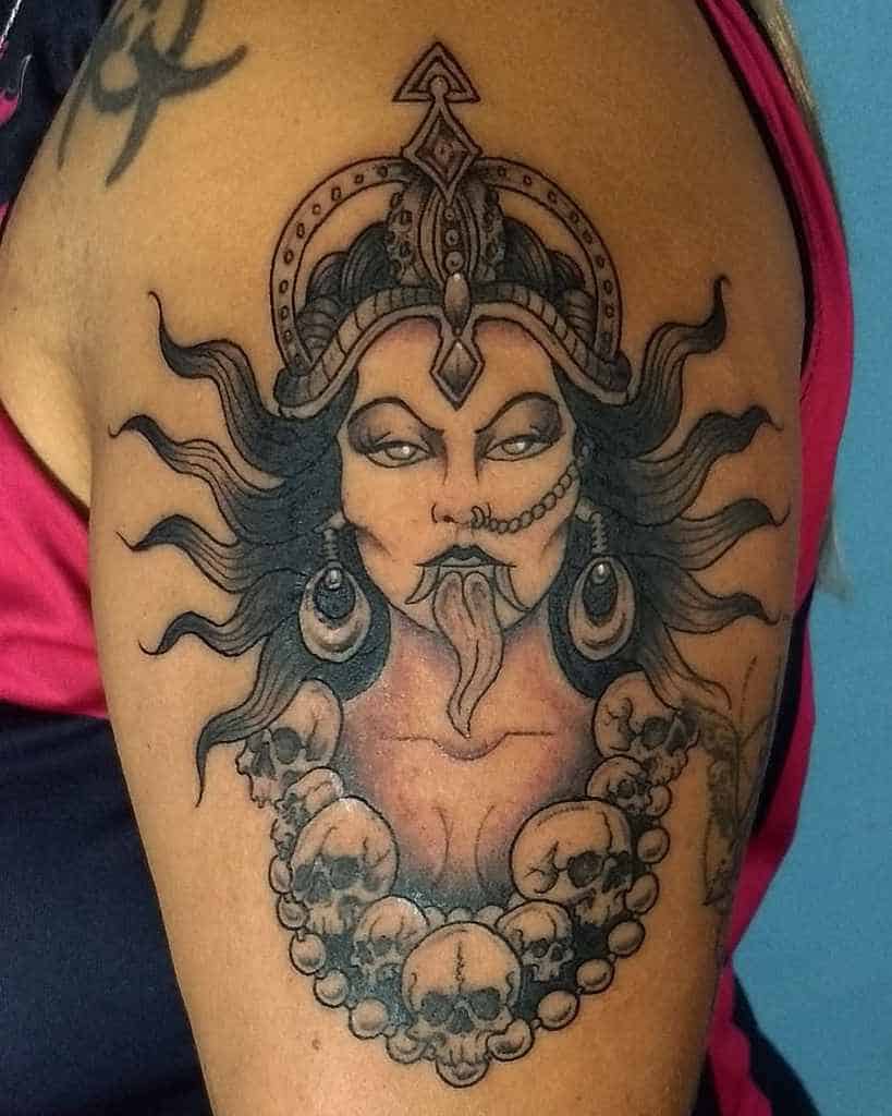 Black and grey Kali tattoo, featuring the goddess with a fierce expression, flowing hair, ornate headdress, extended tongue, and a skull necklace