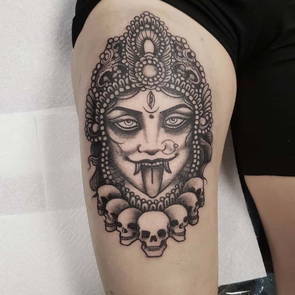 Black and grey Kali tattoo, depicting the fierce goddess with a jeweled headdress, third eye, extended tongue, fangs, and a skull necklace