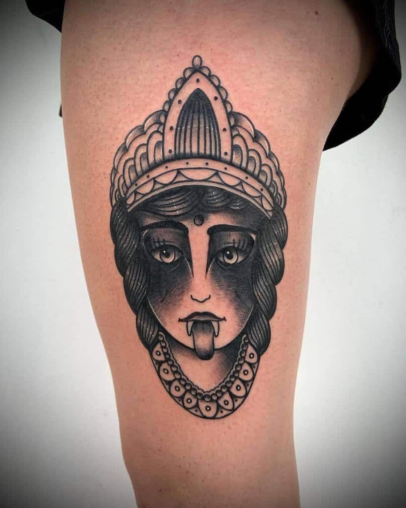 Black and grey Kali tattoo, featuring the goddess with an ornate headdress, third eye, fangs, extended tongue, and beaded necklace