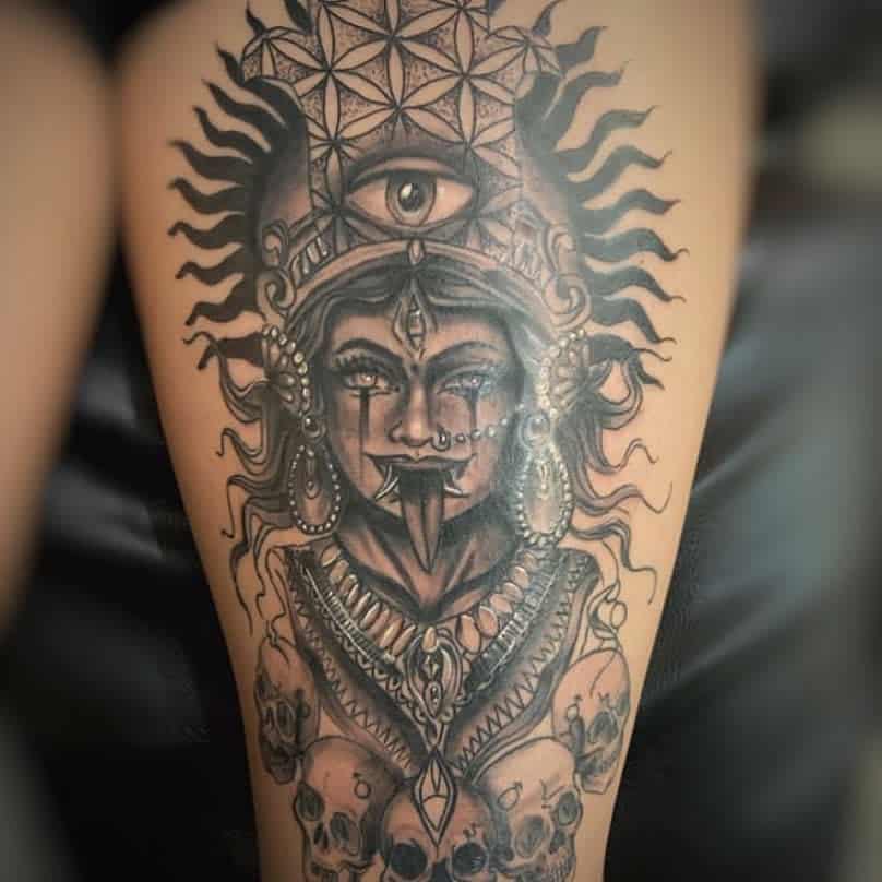 Black and grey Kali tattoo, featuring the fierce goddess with an ornate headdress, a large third eye, flowing hair, extended tongue, and a skull-adorned necklace