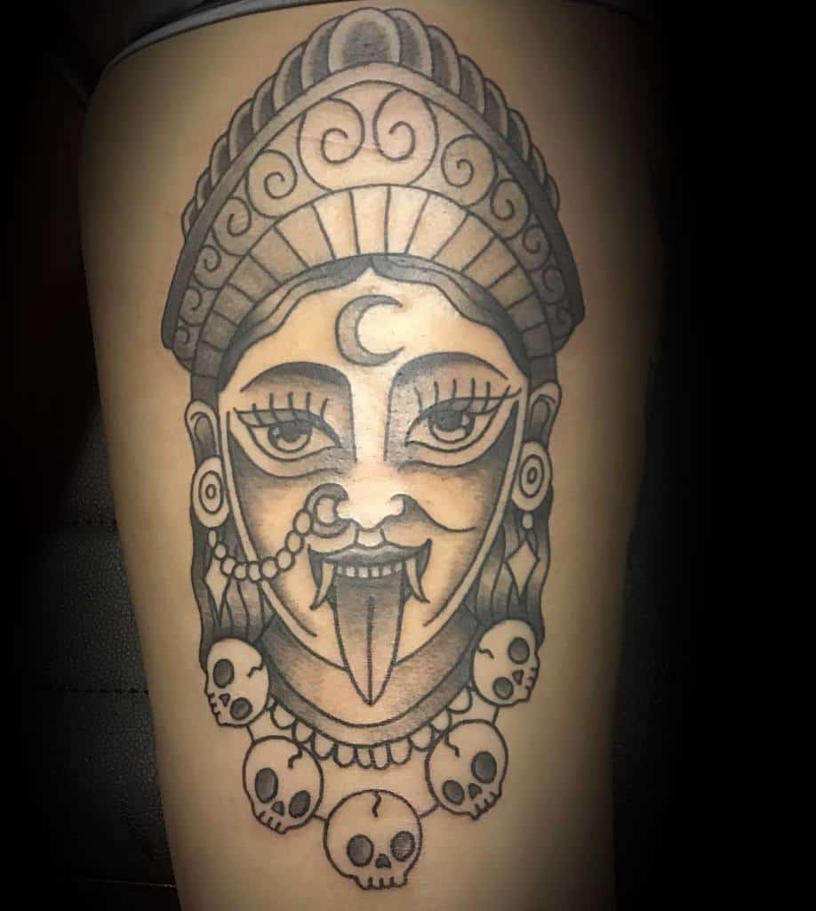 Black and grey Kali tattoo, featuring a bold, stylized goddess with an ornate headdress, crescent moon symbol, extended tongue, and a skull necklace