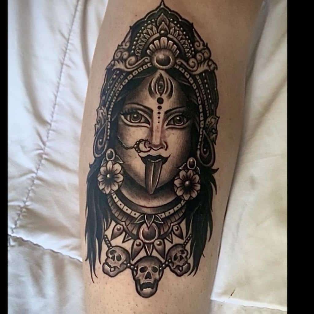 Black and grey Kali tattoo, featuring the goddess with an ornate headdress, third eye, extended tongue, floral earrings, and a skull necklace