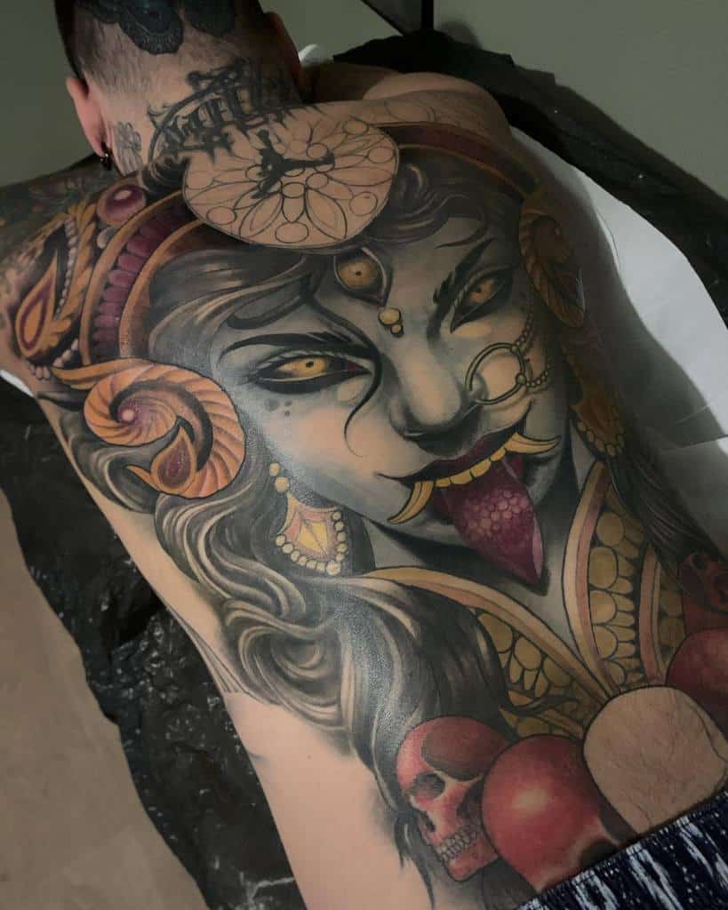 Kali tattoo featuring a fierce, golden-eyed goddess with an ornate headdress, extended tongue, intricate jewelry, and skull-adorned details