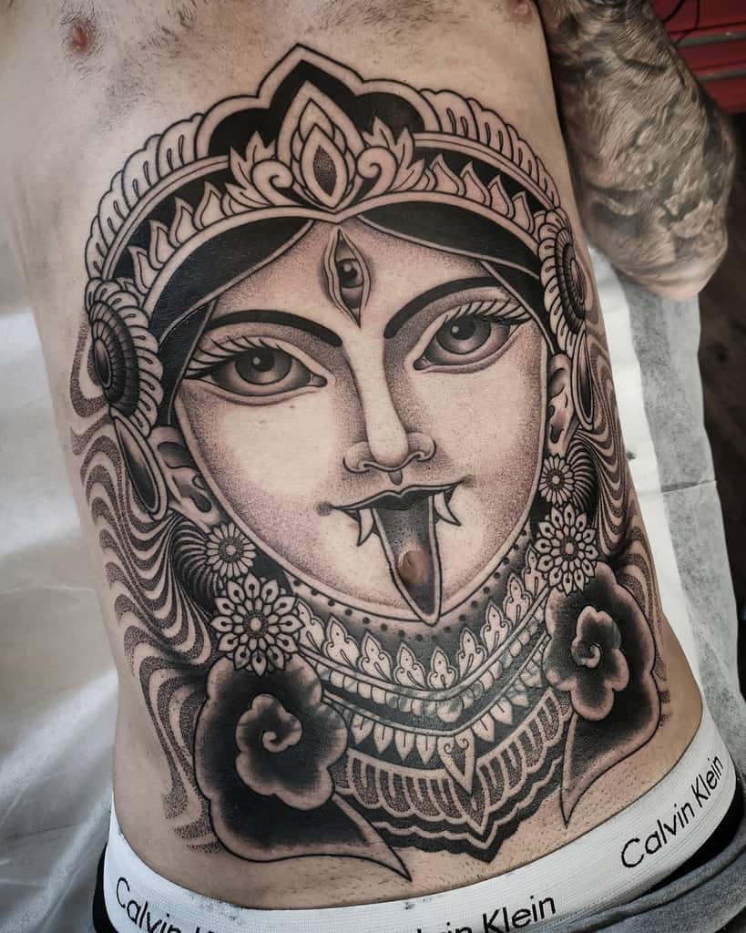 Detailed black and gray tattoo of Kali with a third eye, intricate floral patterns, and a fierce expression