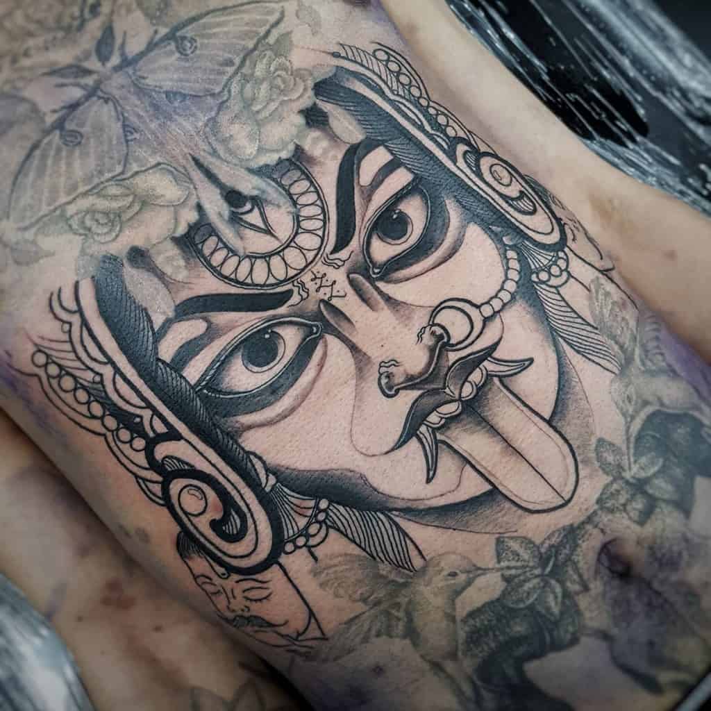 Black and gray tattoo of Kali with a third eye, sticking tongue, surrounded by floral patterns