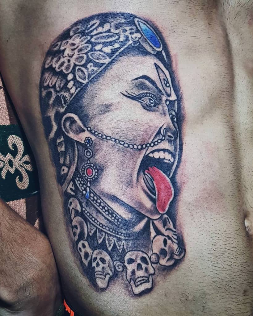 Tattoo of Kali with a third eye, fierce expression, tongue out, adorned with skulls around her neck and intricate details on her head