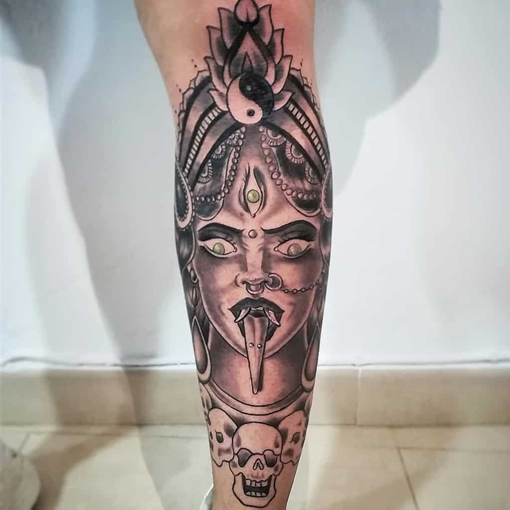 Black and gray tattoo of Kali with a third eye, tongue out, surrounded by skulls and intricate patterns