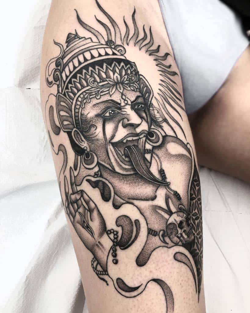 Black and gray tattoo of Kali with a third eye, sticking out her tongue, surrounded by swirling patterns and skulls