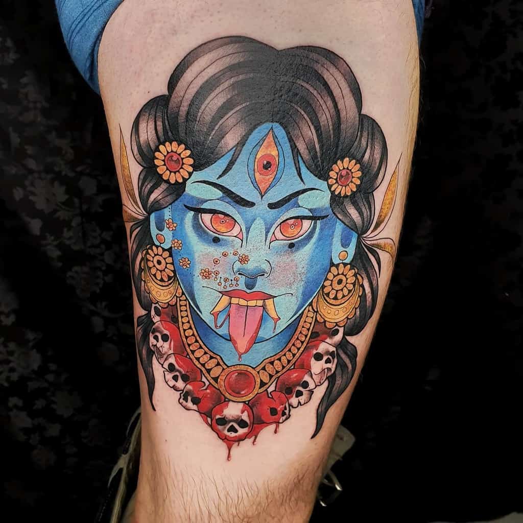 Vibrant tattoo of Kali with blue skin, a third eye, sticking out her tongue, adorned with skulls and floral patterns