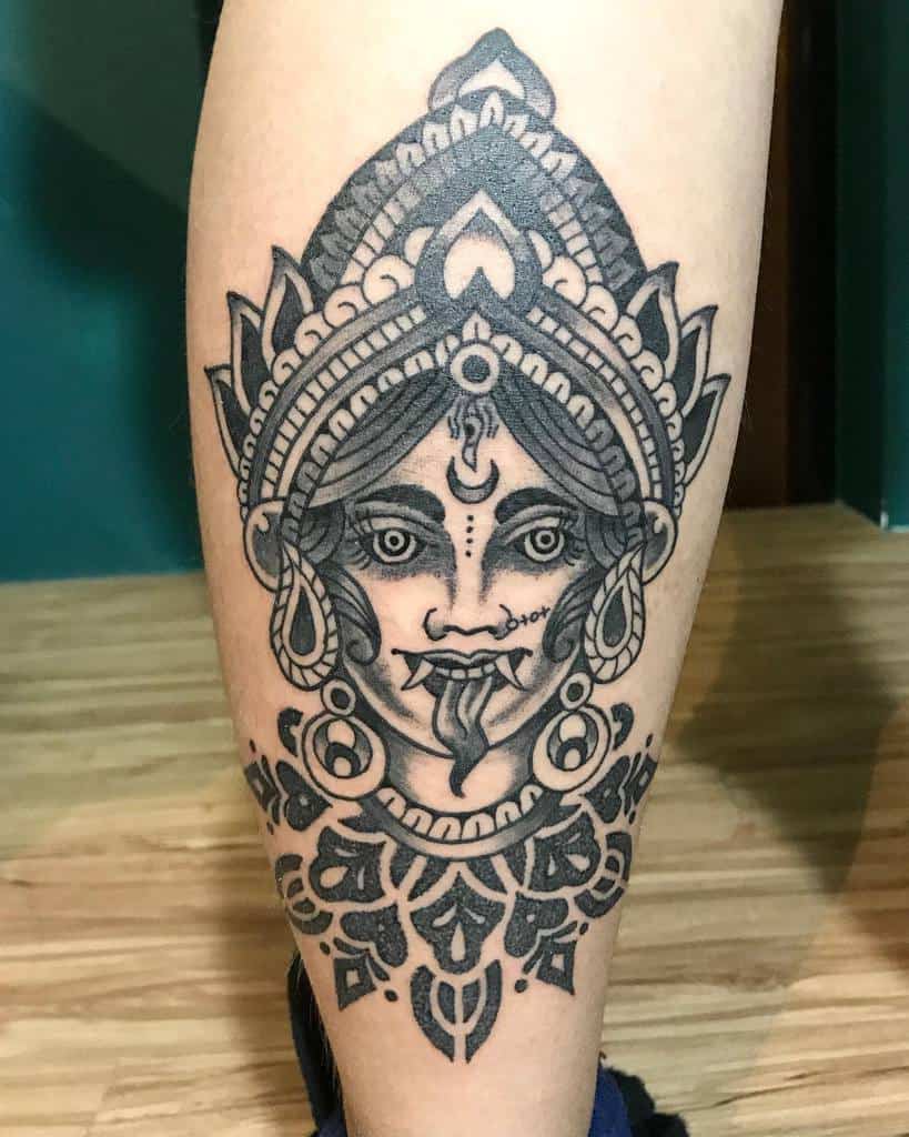 Black and gray tattoo of Kali with a third eye, tongue sticking out, intricate headdress, and ornate patterns
