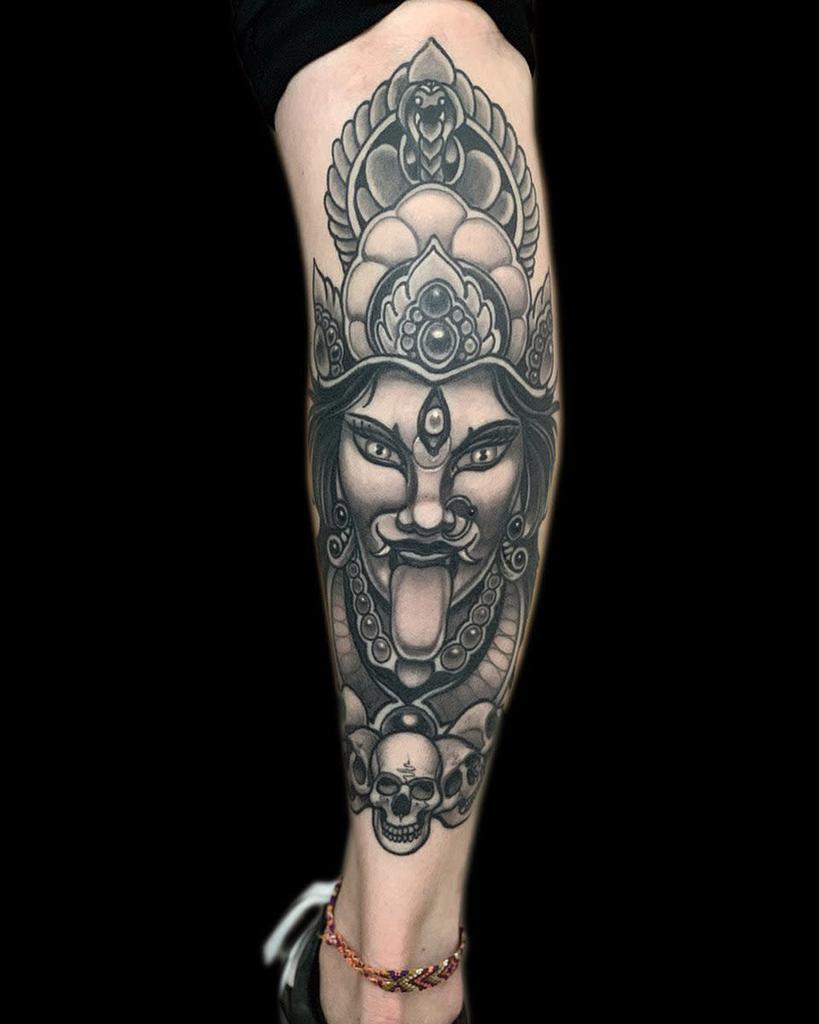 Black and gray tattoo of Kali with a third eye, sticking out her tongue, adorned with a detailed headdress and skulls