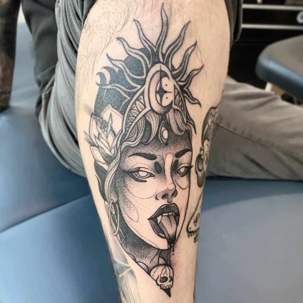 Black and gray tattoo of Kali with a third eye, tongue out, a sun-shaped headdress, and a skull