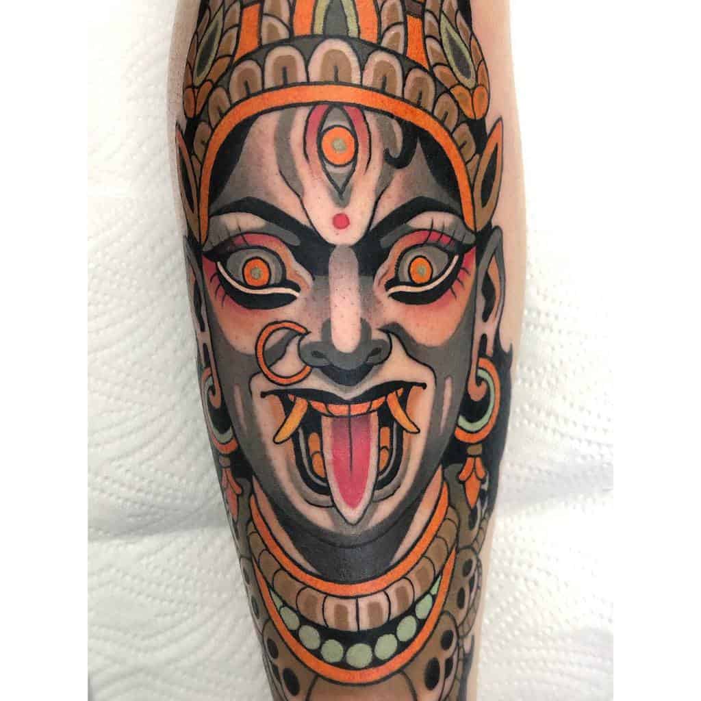 Colorful tattoo of Kali with a third eye, sticking out her tongue, and adorned with intricate jewelry and vibrant patterns
