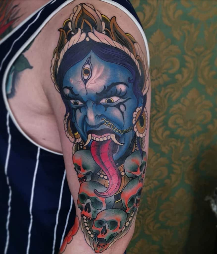 Colorful tattoo of Kali with a third eye, sticking out her tongue, adorned with skulls and intricate details
