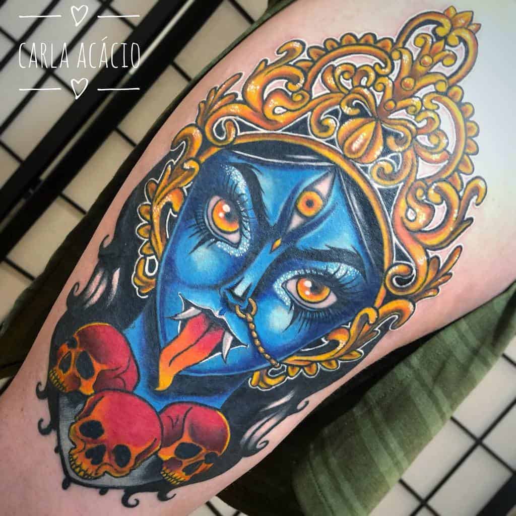 Colorful tattoo of Kali with blue skin, a third eye, sticking out her tongue, wearing an ornate crown, and adorned with skulls