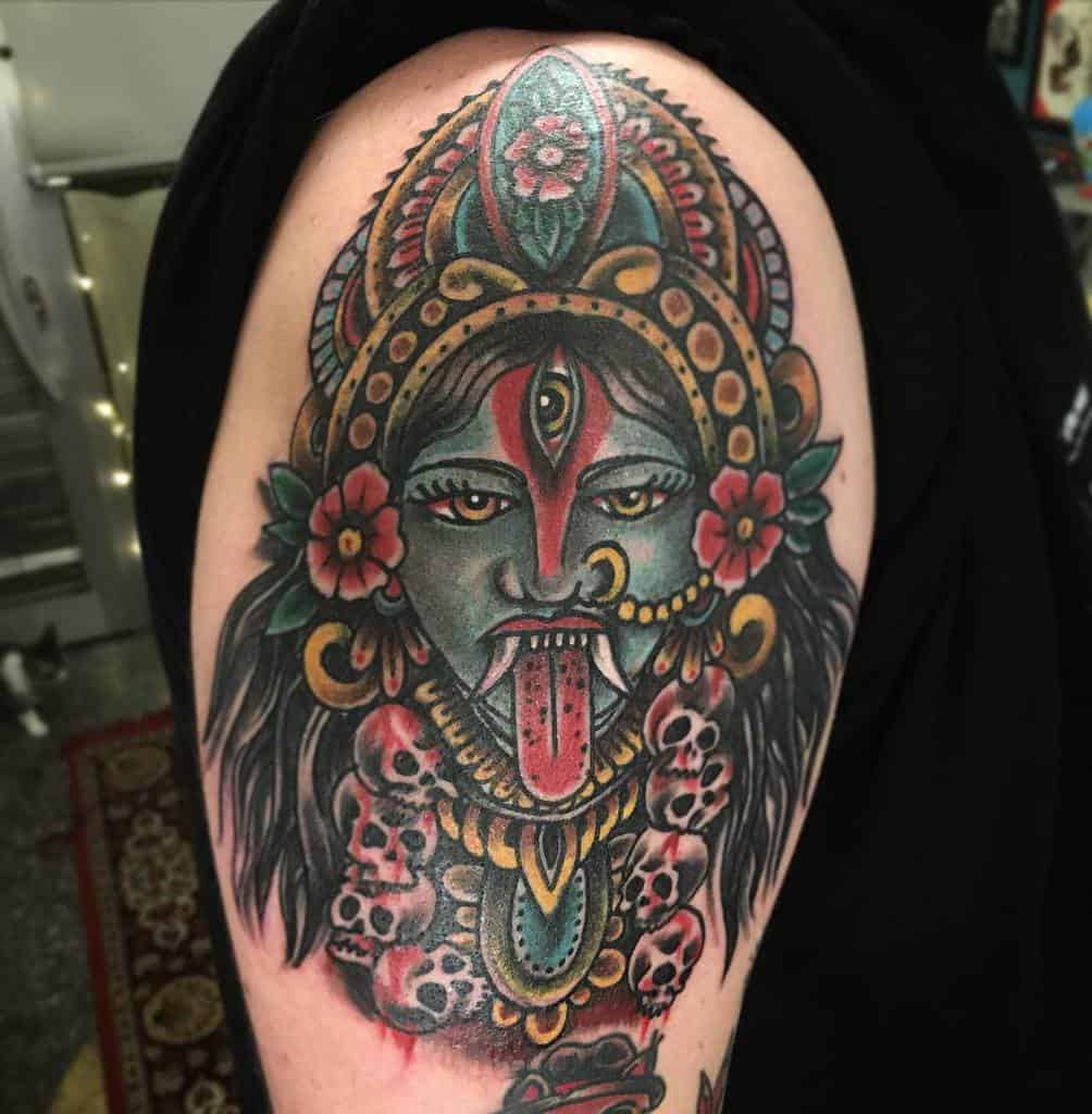 Colorful tattoo of Kali with a third eye, tongue out, adorned with flowers and skulls, and detailed ornamental patterns