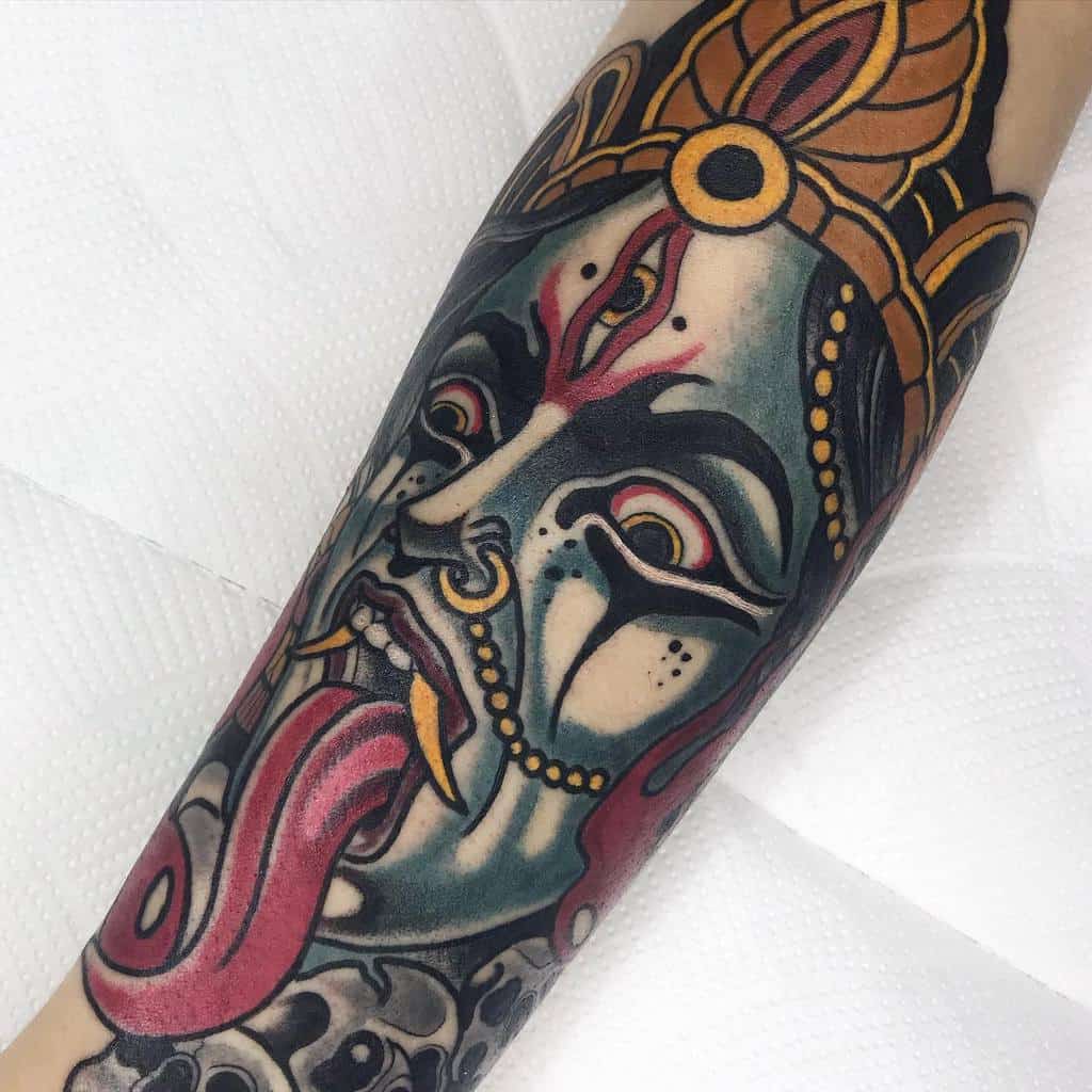 Vibrant tattoo of Kali with a third eye, sticking out her tongue, adorned with intricate patterns, and skulls
