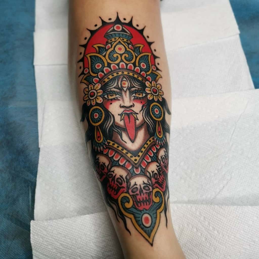 Colorful tattoo of Kali with a third eye, sticking out her tongue, wearing a vibrant headdress, and adorned with flowers and skulls