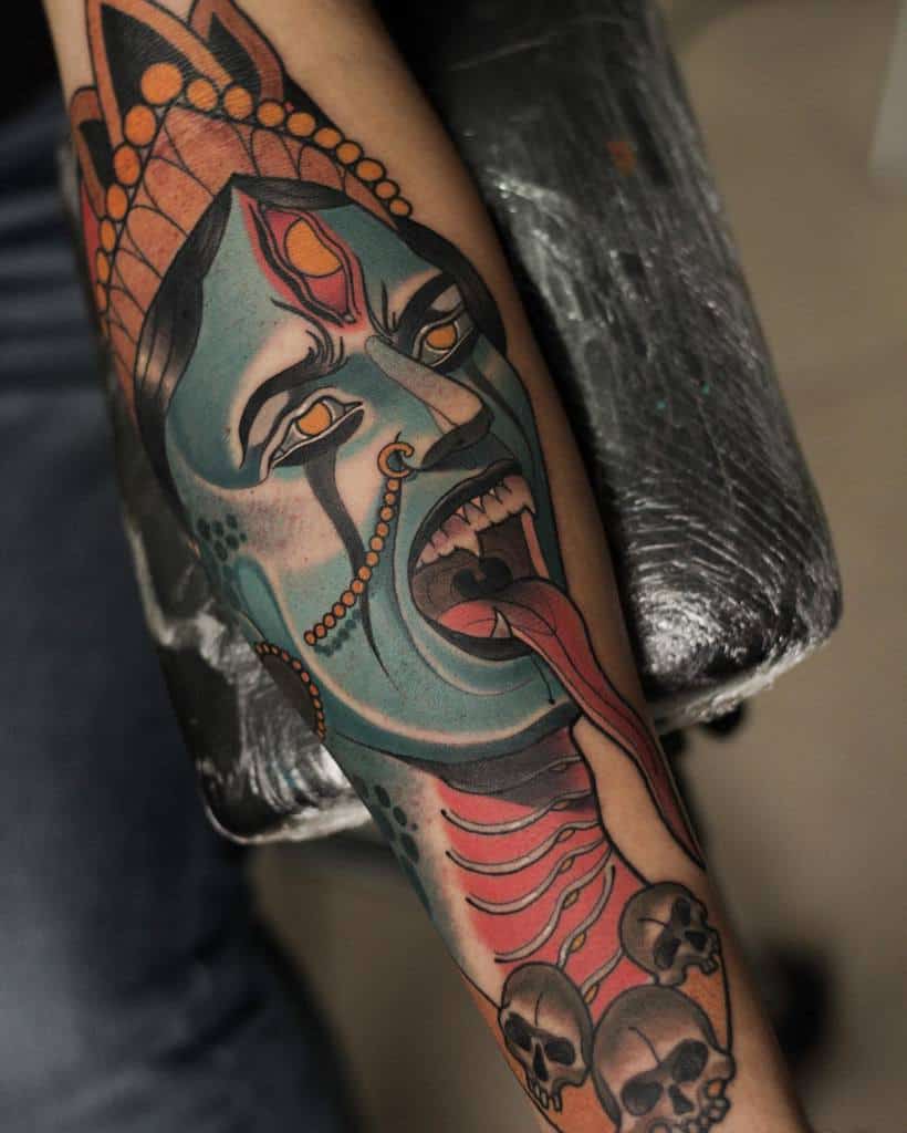 Colorful tattoo of Kali with a third eye, sticking out her tongue, wearing a crown, adorned with skulls, and intricate details