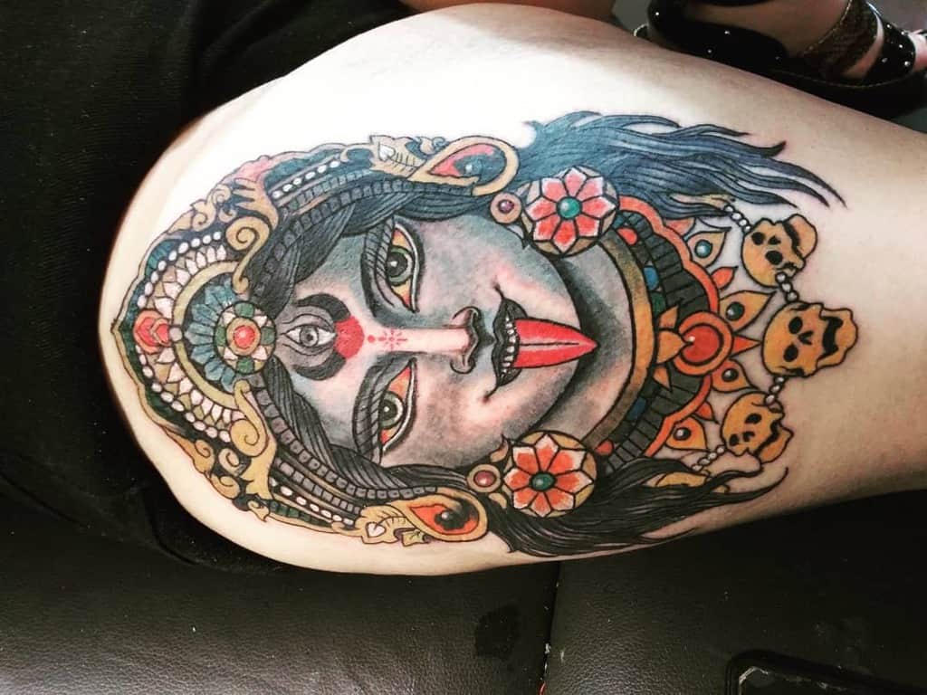 Colorful tattoo of Kali with a third eye, sticking out her tongue, adorned with skulls, floral patterns, and an ornate headdress