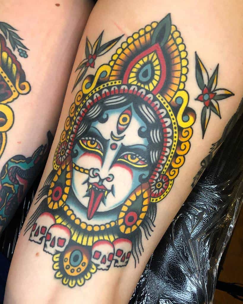 Colorful tattoo of Kali with a third eye, sticking out her tongue, wearing an ornate headdress, adorned with skulls, and surrounded by floral accents