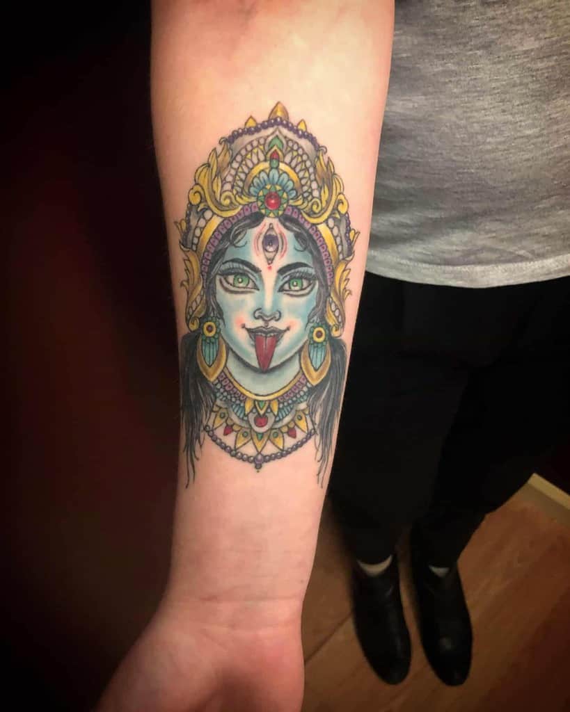 Colorful tattoo of Kali with a third eye, sticking out her tongue, adorned with a vibrant headdress and intricate jewelry