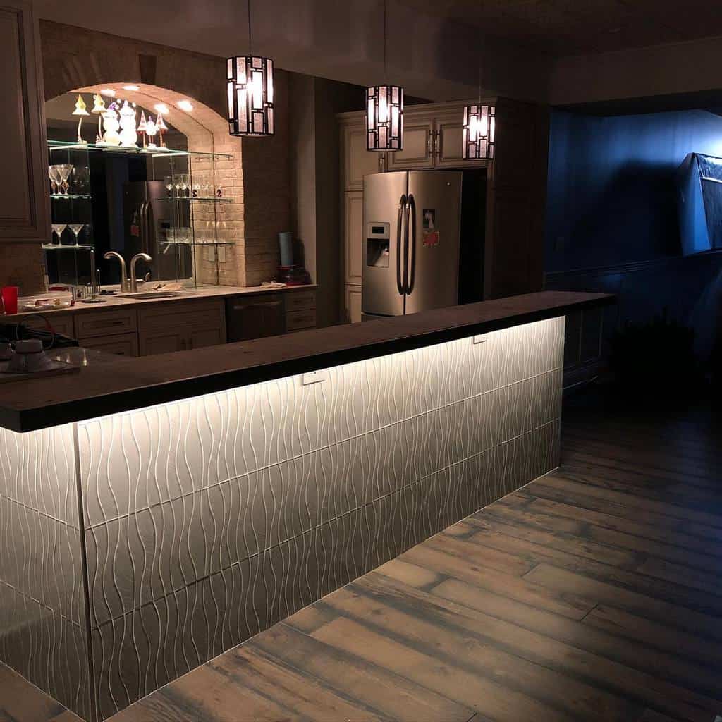 Modern basement bar with backlit tile design, pendant lights, and glass display shelves.