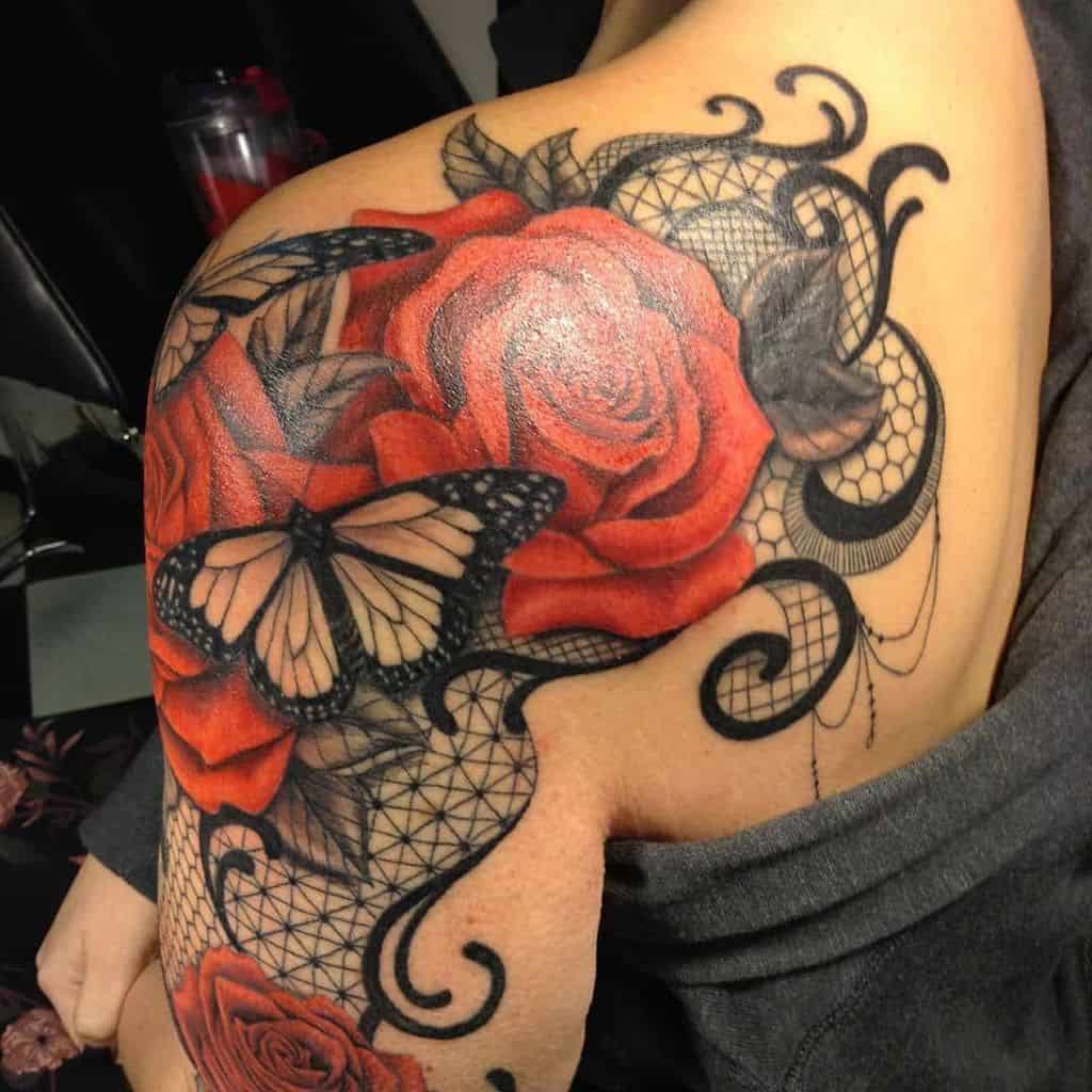 Bold shoulder tattoo featuring vibrant red roses, a detailed monarch butterfly, and intricate black lace patterns with ornamental swirls.