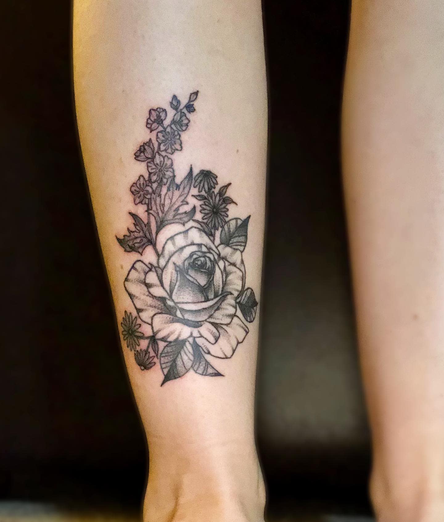 Forearm tattoo of a detailed rose with larkspur flowers and leaves extending upwards