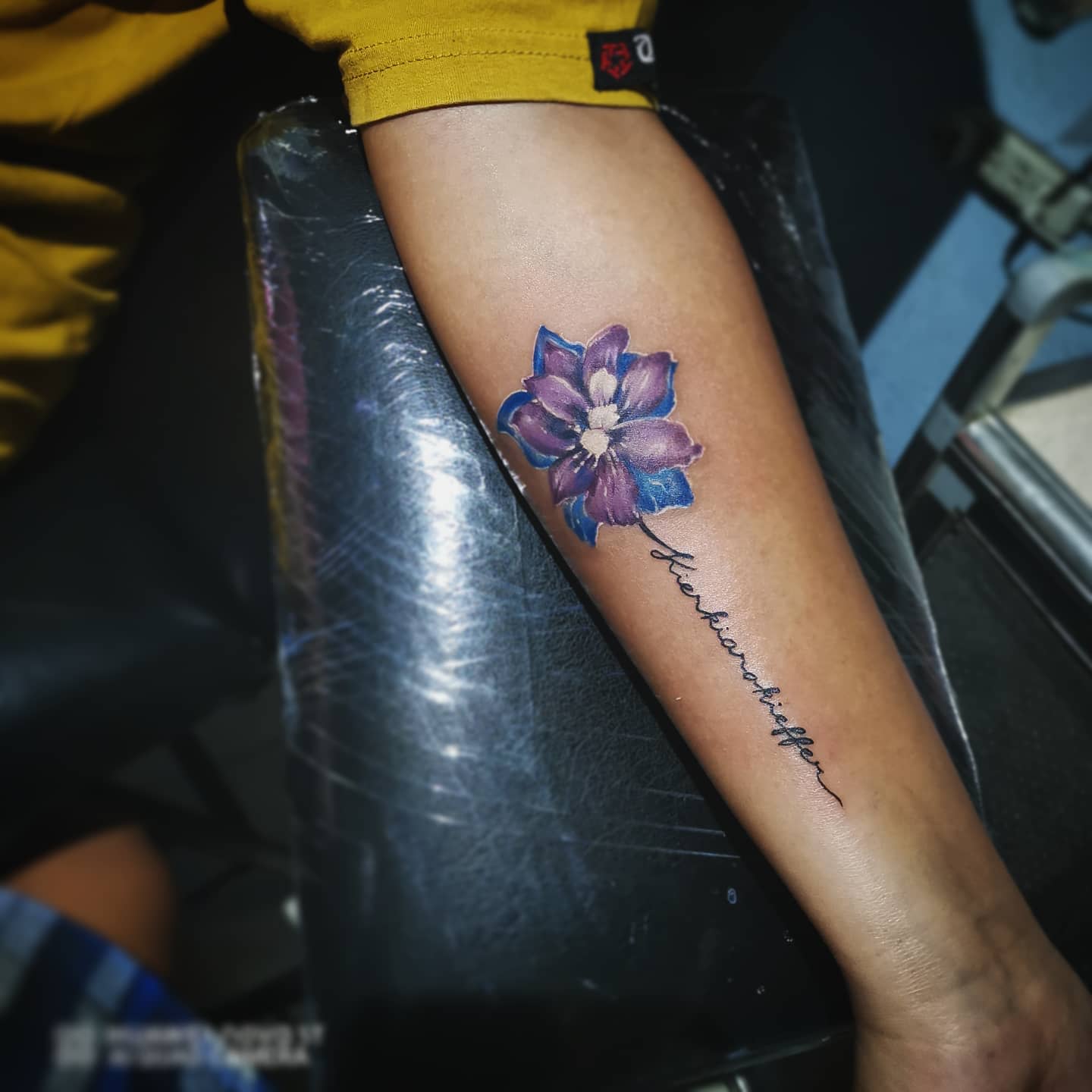 Forearm tattoo featuring a purple flower with a cursive word below it