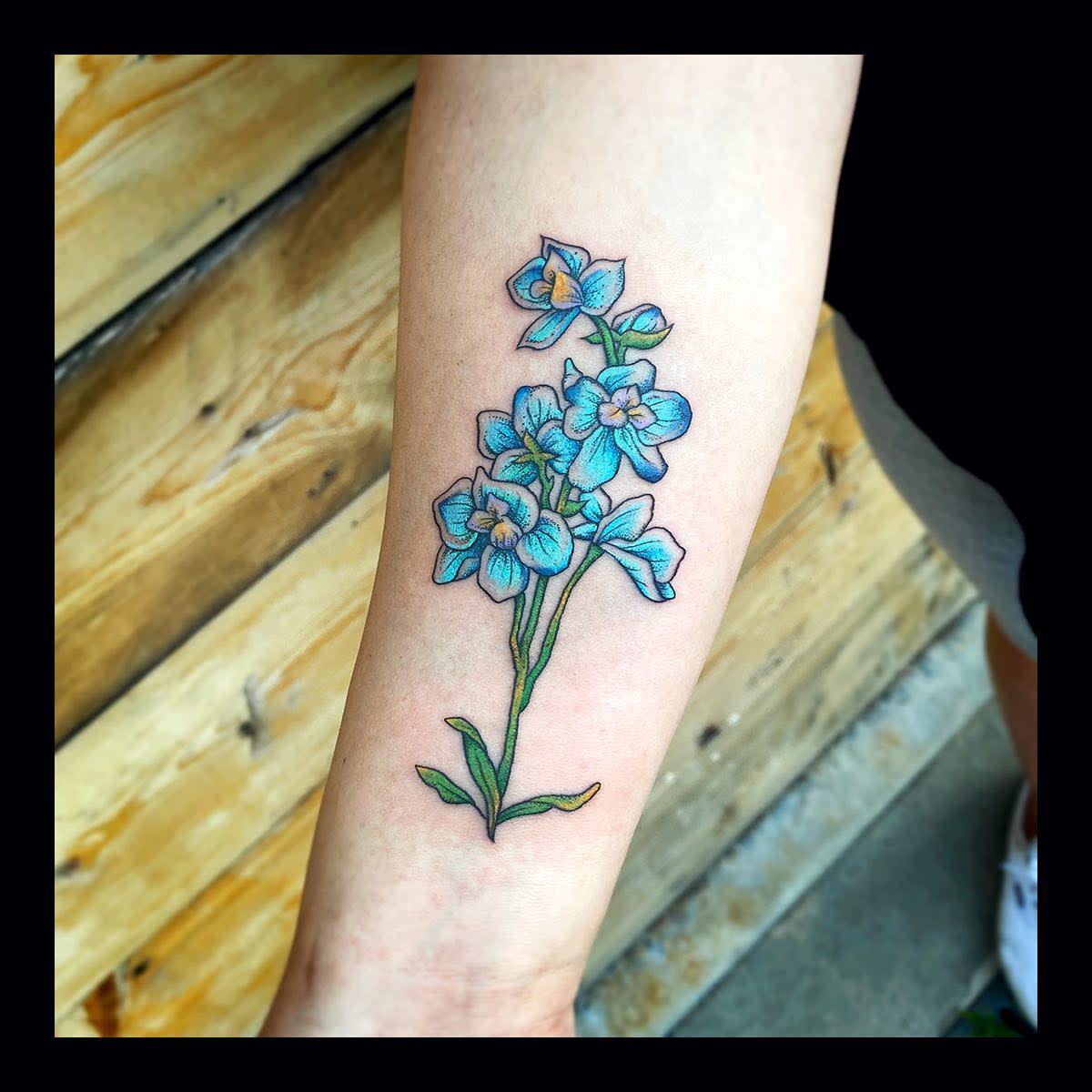 Forearm tattoo of a blue flower bouquet with green stems against a wooden background