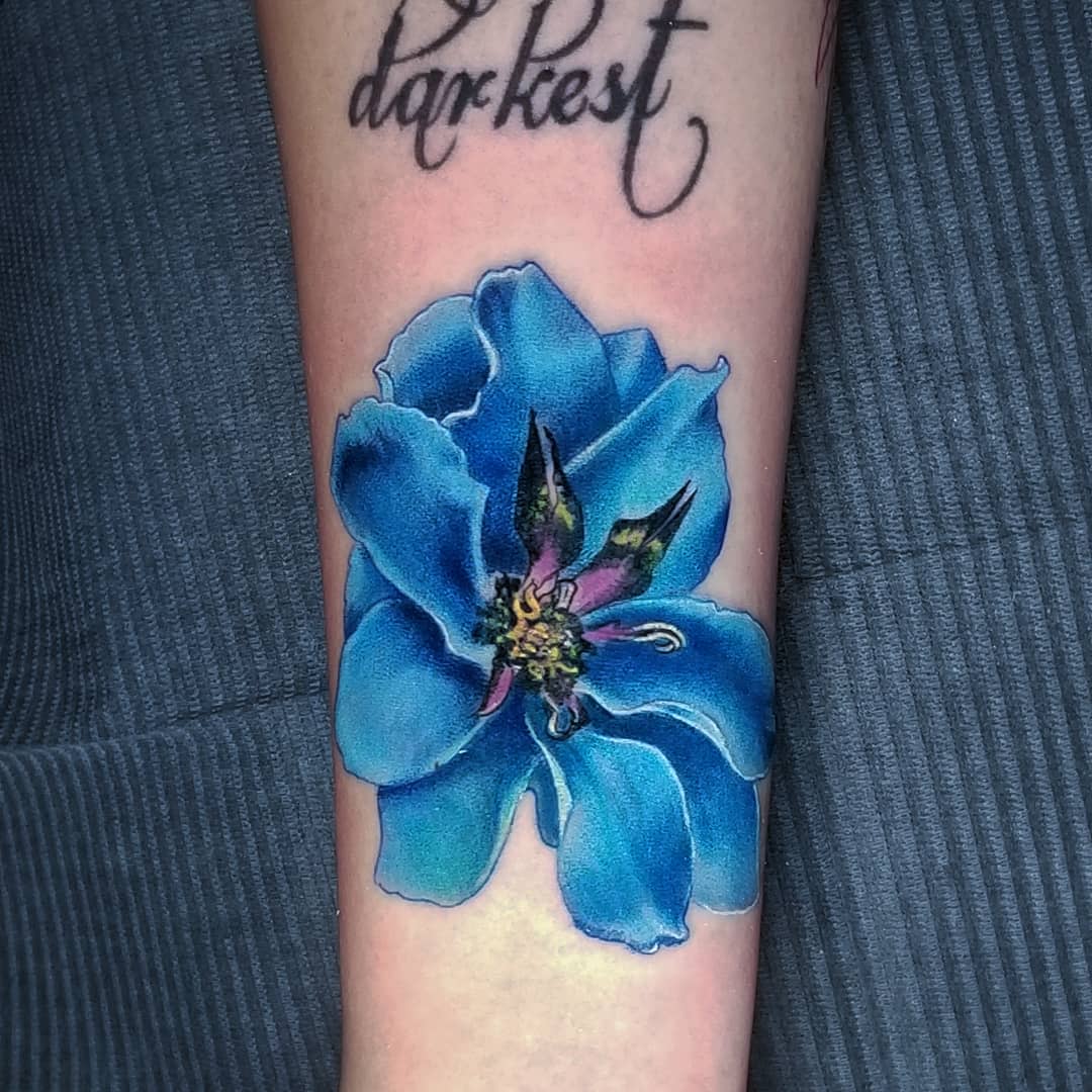 Tattoo of a large, detailed blue flower on a forearm with the word "darkest" above it