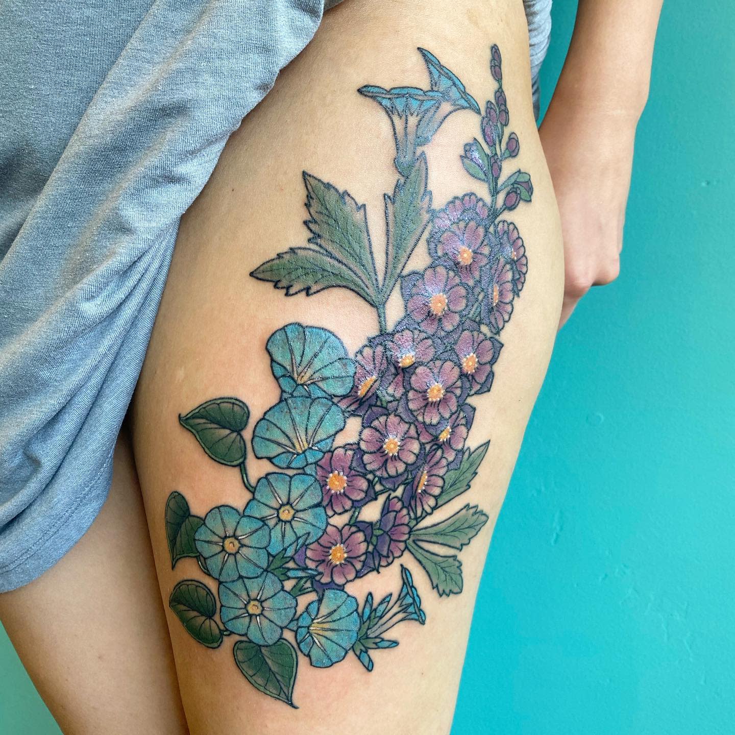 Thigh tattoo featuring blue and purple flowers with green leaves