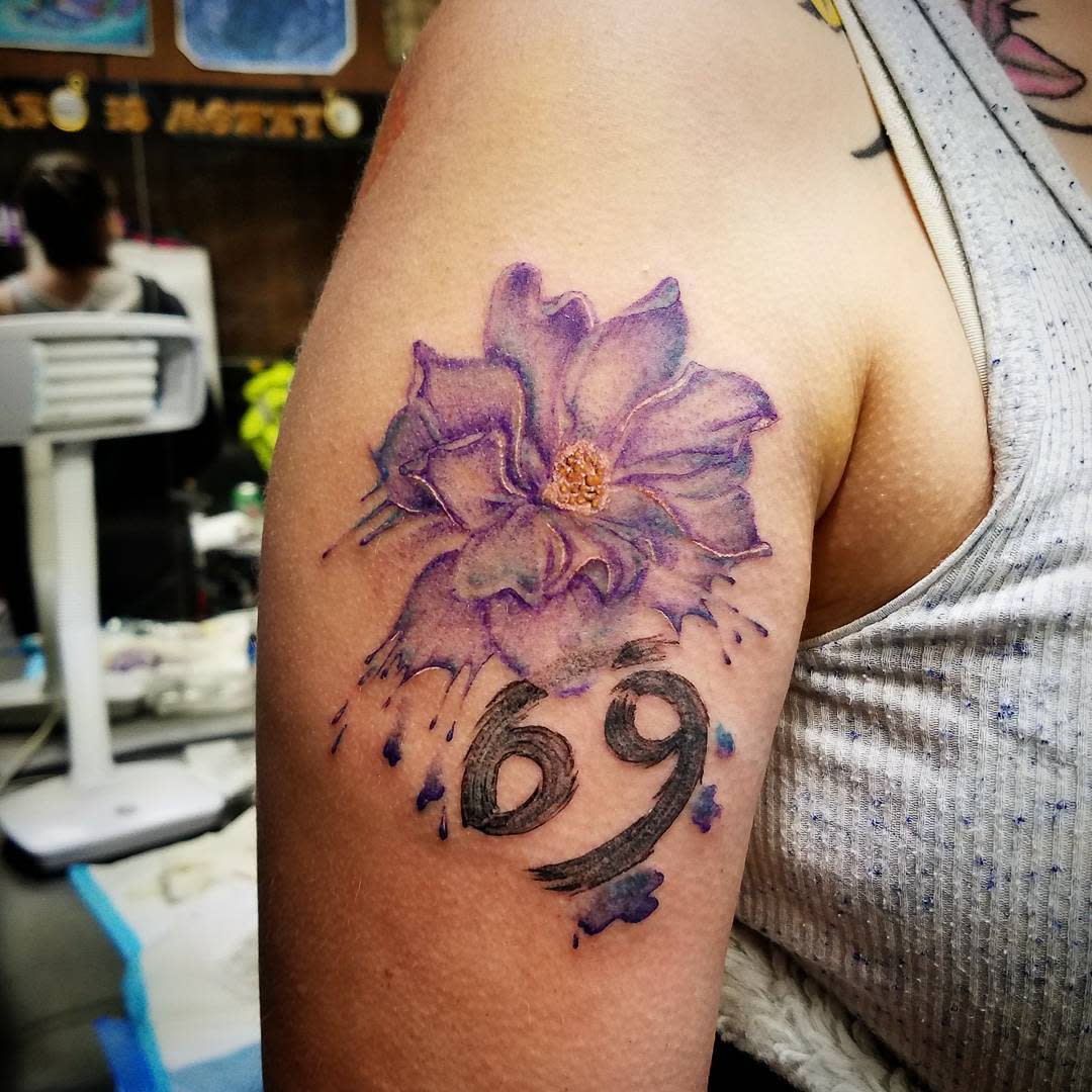 Purple flower tattoo with the number 69 in black ink on an individual's upper arm