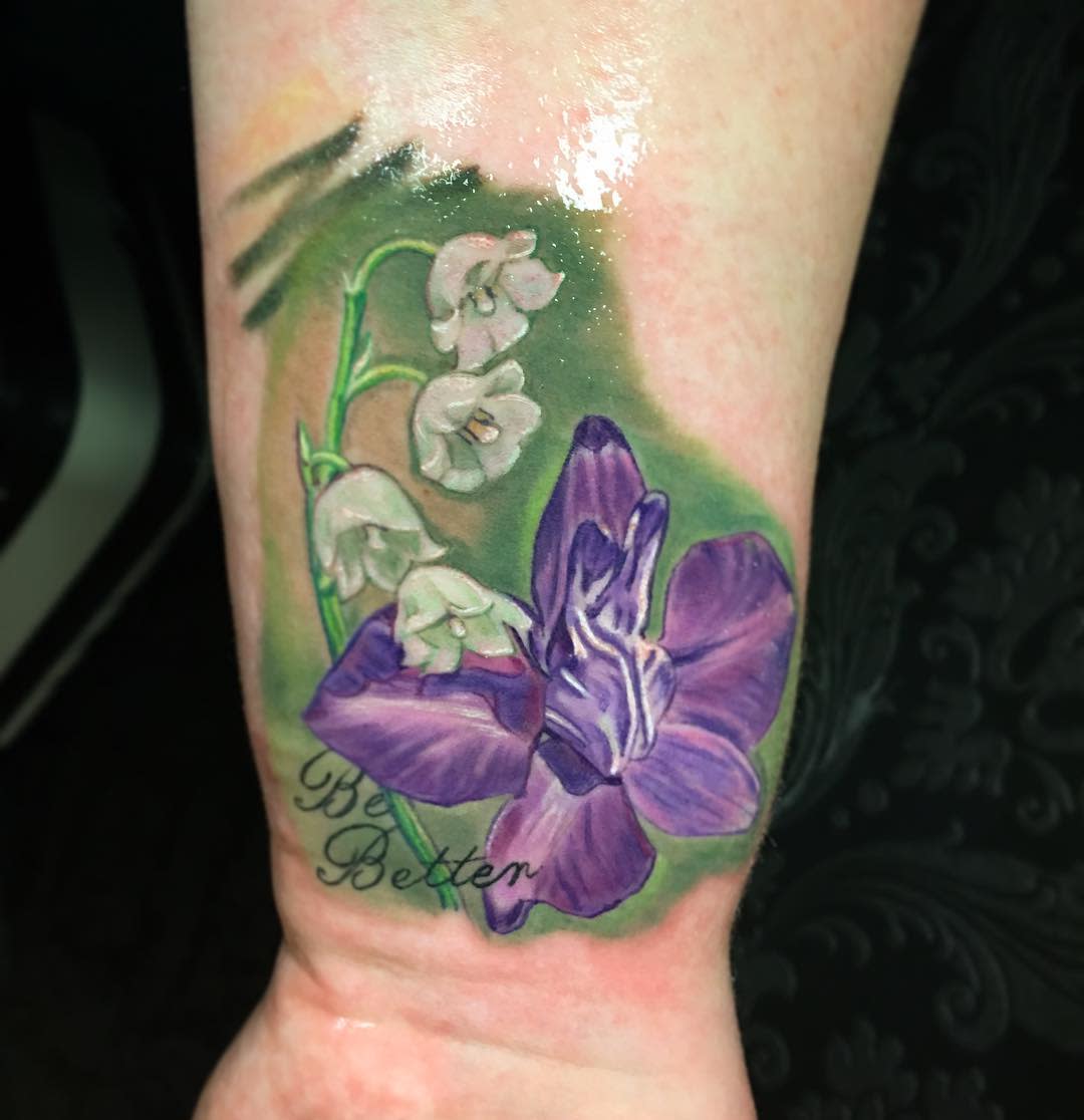 Tattoo of purple and white flowers on forearm with "Be Better" text below the flowers