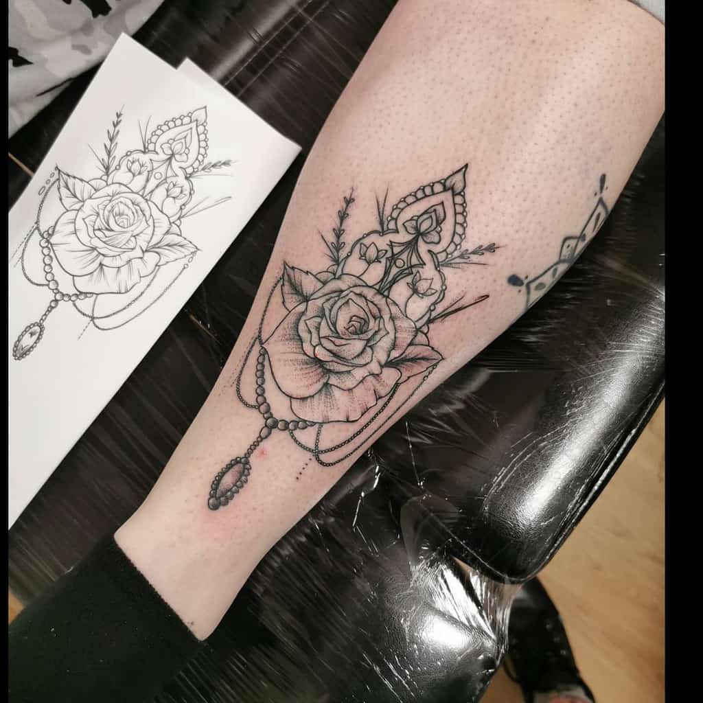 Black and grey leg tattoo featuring a detailed rose, ornamental filigree, chandelier-style beadwork, and a teardrop jewel accent