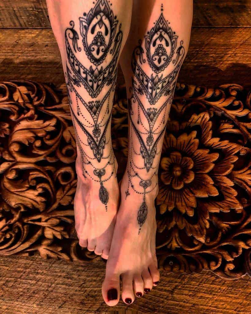 Elegant symmetrical leg tattoos featuring intricate ornamental patterns, chandelier-style beadwork, and teardrop accents