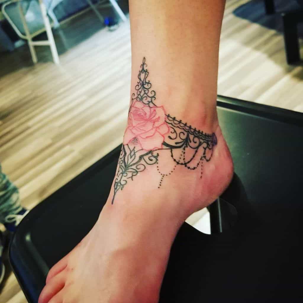 Elegant ankle tattoo featuring a black lace garter design with a delicate pink rose, intricate filigree, and chandelier-style beadwork